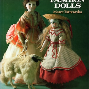 May include: A book cover titled "Fashion Dolls" by Maree Tarnowska. The image features two antique dolls in period clothing, a small dog figurine, and text. The dolls are dressed in elaborate gowns with hats. The background is a solid green.