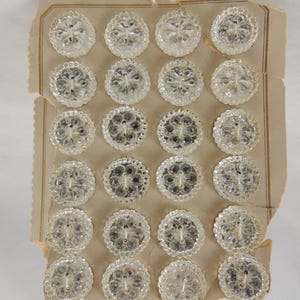 May include: A collection of clear, round buttons with a floral design, arranged on a vintage cardboard backing. Each button features a scalloped edge and a central flower motif. The buttons are made of a transparent material, allowing light to pass through.