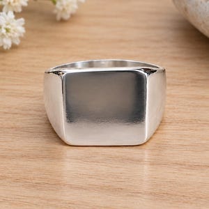 May include: A polished silver signet ring featuring a smooth, flat, square-shaped face. The band is solid and metallic with a reflective surface, showcasing a clean, unadorned design that highlights the bright, lustrous finish of the silver metal.