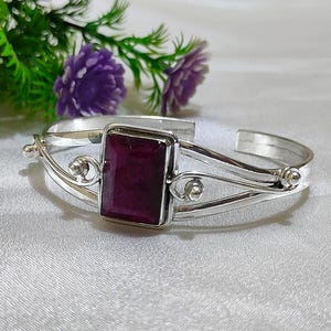 May include: A silver cuff bracelet featuring a large, rectangular, deep red gemstone. The bracelet has decorative swirls and small silver balls. The background includes purple flowers and green foliage.