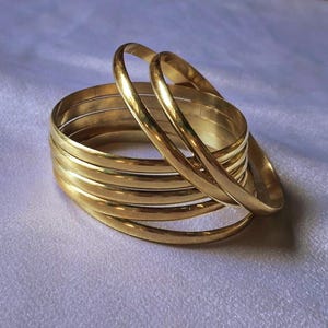 Peut inclure: Plusieurs bracelets dorés, certains empilés et d'autres séparés, reposent sur une surface blanche texturée. Les bracelets ont une finition lisse et polie, reflétant la lumière. Les bijoux sont probablement destinés à être portés au poignet.
