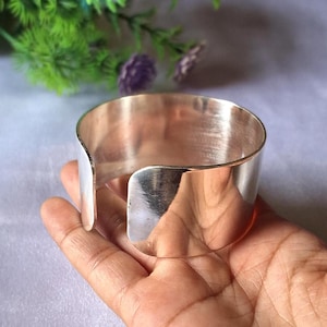 May include: A wide, polished silver cuff bracelet. The open design allows for easy wear. The bracelet has a smooth, reflective surface, and is approximately 5 cm wide. The cuff is held in a hand, with a blurred background of greenery and purple flowers.