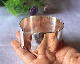 May include: A wide, polished silver cuff bracelet. The open-ended design allows for easy wear. The bracelet has a smooth, reflective surface, and is approximately 2 inches wide. The cuff is held in a hand, with a blurred background of greenery and purple flowers.