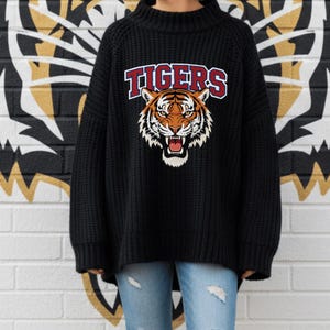 May include: A person wearing a black oversized sweater with the word "TIGERS" and a tiger graphic on the front, paired with ripped blue jeans. The background features a white brick wall with a large tiger head graphic.