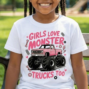 Può includere: Una bambina indossa una maglietta bianca con una grafica di monster truck rosa e la scritta "GIRLS LOVE MONSTER TRUCKS TOO". La bambina sorride, seduta su una panchina in un ambiente verde.