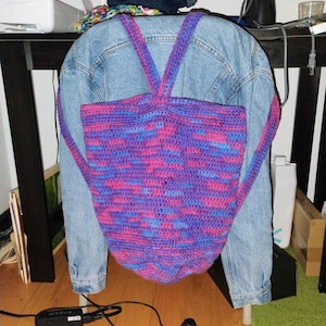 May include: A handmade crochet backpack in shades of purple, pink, and blue. The backpack has adjustable straps and a rounded shape. It is hanging on a denim jacket.