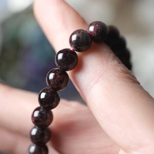 Puede incluir: Una pulsera de cuentas de color rojo oscuro. Las cuentas son redondas y tienen un acabado brillante. La pulsera está ensartada en un cordón fino de color rosa. La pulsera se muestra en un dedo.