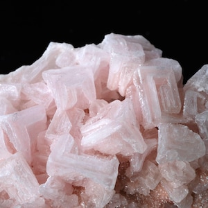 Puede incluir: Primer plano de un grupo de cristales de halita de color rosa claro. Los cristales tienen una estructura cúbica y un aspecto translúcido, con una textura ligeramente rugosa. El fondo es negro, resaltando el color y la forma del mineral.