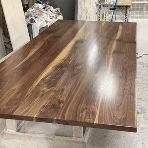 May include: A large, rectangular wooden table with a polished, glossy finish. The tabletop is made of dark brown wood with lighter brown streaks, showcasing the natural grain. The table is supported by a light-colored base.