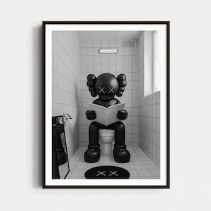 Op de afbeelding: Een ingelijste muurkunstprint met een zwart stripfiguur met X-vormige ogen die op een wit toilet in een betegelde badkamer zit en een krant leest. Een zwart rond tapijt met X-vormige ogen ligt op de vloer voor het toilet.