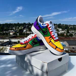 May include: A pair of custom-painted Air Force 1 sneakers. The shoes feature a vibrant, multi-colored design with a white sole and laces. The color scheme includes yellow, orange, green, blue, and purple, with black accents. The shoes are displayed on a white box.