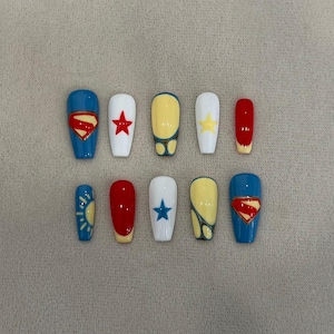 Pode incluir: Um conjunto de dez unhas postiças com vários designs, incluindo o logótipo do Super-Homem, estrelas vermelhas e azuis, padrões amarelos e azuis e detalhes em vermelho sólido. As unhas estão dispostas em duas filas de cinco com acabamento brilhante.