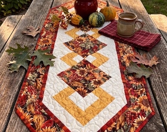 May include: A quilted table runner with a fall theme. The runner features a central design of white fabric with yellow geometric shapes and diamond-shaped floral patterns. The edges are bordered with a floral print of sunflowers and autumn leaves. A mug of coffee, pumpkins, and a vase of sunflowers are on the table.