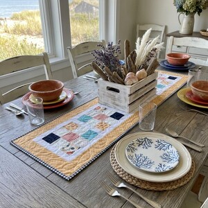 May include: A dining table set with a quilted table runner featuring a patchwork pattern of blue, orange, and white squares. A white wooden crate centerpiece holds dried lavender, driftwood, and shells. Place settings include ceramic plates.