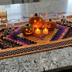 May include: A rectangular quilted table runner featuring a geometric pattern in shades of orange, purple, and black. The fabric displays motifs of bats and pumpkins. It is decorated with two jack-o'-lanterns, two small LED candles, and string lights.
