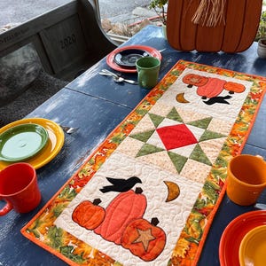 May include: A quilted table runner with a fall theme. The runner features orange pumpkins, a black crow, and a starburst design. The border is decorated with fall leaves. The runner is on a blue table with colorful plates and mugs.