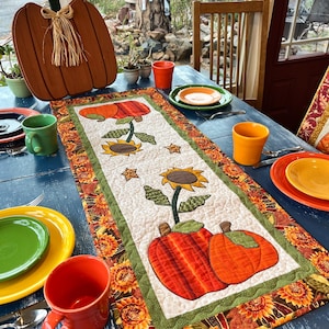 May include: A fall-themed table setting with a blue table, featuring a quilted table runner with pumpkin and sunflower designs. The table is set with colorful plates, mugs, and a wooden pumpkin decoration.