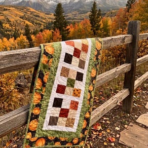 May include: A handmade fall-themed table runner draped over a wooden fence. The runner features a patchwork design with squares of brown, orange, and green fabrics. The border is green with pumpkin patterns. The background shows a scenic autumn landscape.