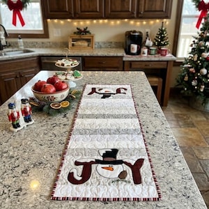 May include: A rectangular quilted table runner featuring a white background with a swirling pattern and two snowman motifs. Each snowman is centered between the letters J and Y, spelling the word JOY in dark red fabric with a black top hat.