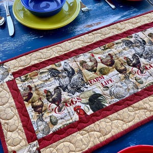 May include: A quilted table runner with a farm theme. The center features roosters, chickens, and eggs with the words "Farm Life" and "Eggs". The runner has a red border and a beige honeycomb pattern on the sides. The table is set with blue, yellow, and red dishes.