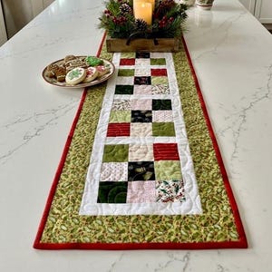May include: A rectangular quilted table runner featuring a patchwork center of red, green, white, and black squares with various holiday patterns. The runner has a wide border with a green holly leaf print and a thin red edge trim.