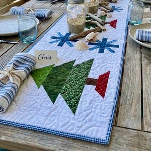 May include: A white quilted table runner featuring appliqued green fabric Christmas trees, red trunks, and blue snowflake patterns. The runner has a blue gingham border and is decorated with seashells, driftwood, and a place card reading Chris.
