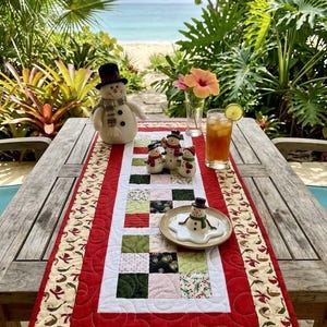 May include: A rectangular quilted table runner featuring a central patchwork grid of red, green, white, and black squares with holiday patterns, bordered by a solid red band and an outer edge of cream fabric decorated with small red and green motifs.