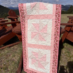 May include: A handmade pink and white quilt with a floral border. The quilt features a pinwheel design in the center, with a red plaid pattern in between the pinwheels. The quilt is displayed outdoors.