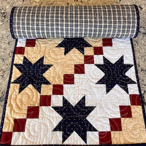 May include: A quilted table runner featuring a pattern of dark blue stars with white dots on a background of cream and white fabric squares. The design includes red square accents and a dark blue plaid border with continuous swirl quilting.