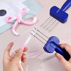 May include: Two blue knitting tools with metal prongs are shown, alongside a partially completed knitted piece in white and pink yarn. A finished yarn bow in pink and white is also visible. The tools are for knitting.