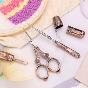 Könnte beinhalten: Ein Set aus kupferfarbenen Nähwerkzeugen, bestehend aus einer Schere mit floralen Gravuren, einer spitzen Ahle mit passendem Griff, einem Fingerhut und einer Schutzhülle für die Ahle, alle mit detaillierten geprägten Metallverzierungen.