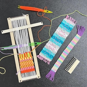 Peut inclure: Un métier à tisser en bois avec des fils de chaîne blancs et un textile partiellement tissé orange, rose et jaune. Comprend une aiguille à tisser verte, du fil orange, une bande tissée bleue et blanche, un ruban multicolore et un peigne en bois.