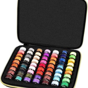 Puede incluir: Un estuche negro con cremallera dorada, abierto para revelar una colección de bobinas de máquina de coser coloridas. Las bobinas están dispuestas en filas, mostrando una variedad de colores de hilo, incluyendo rojo, azul, verde y morado.