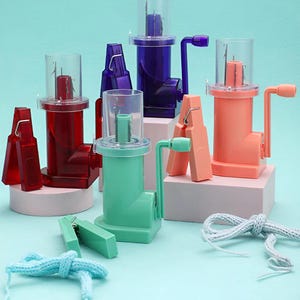 Peut inclure: Plusieurs machines à tricoter colorées en rouge, violet, vert et pêche. Chaque machine est dotée d'un cylindre en plastique transparent et d'une manivelle. Des accessoires et du fil tricoté sont également présents, suggérant un thème artisanal.