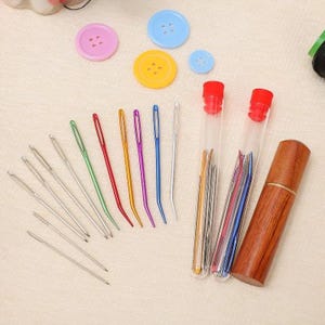 May include: A collection of sewing needles in various colors and sizes, along with colorful buttons. The needles are arranged with a wooden needle case and clear tubes with red caps. The buttons are pink, blue, and yellow.