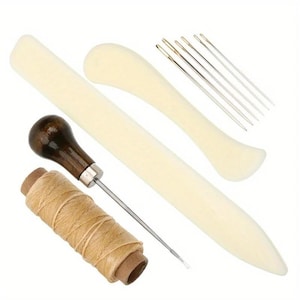 May include: A leather crafting tool set including a long straight bone folder, a curved bone folder, five silver metal needles, a wooden-handled awl with a metal tip, and a spool of tan thread. These tools are arranged together on a white background.