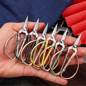 May include: A collection of six small, decorative embroidery scissors with silver blades and ornate handles in various metallic finishes, including silver, black, gold, and iridescent colors. The scissors are held in a hand.