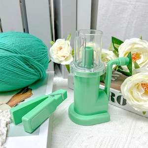 May include: A mint green yarn winder with a clear plastic top and a hand crank. A matching green clothes peg and a ball of teal yarn are also visible. White flowers and a decorative box are in the background.
