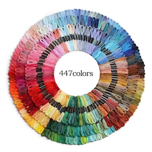 Puede incluir: Una disposición circular de 447 madejas de hilo de bordar, que muestra una amplia gama de colores. El hilo está cuidadosamente organizado en un patrón de arcoíris, que va del rojo y el naranja al verde, el azul y el morado. El texto "447 colors" está en el centro.