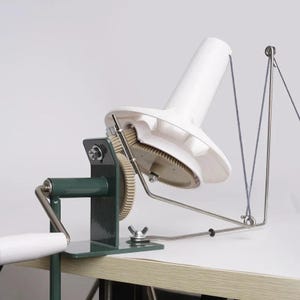 May include: A white and green yarn swift with a hand crank. The yarn swift has a large white cone-shaped top and a metal frame. The base is green and has a hand crank with a white handle. The yarn is a light blue color.
