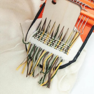 May include: Close-up of a weaving project in progress. A wooden weaving tool is positioned over a small woven piece with a fringe of colourful threads in shades of green, yellow, and brown. The project is set against a white fabric background.
