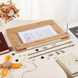 May include: A rectangular bamboo bookbinding press with two vertical threaded metal rods and wing nuts, holding a stack of white paper. Included are two additional long metal rods, several small metal fasteners, and adhesive black circular pads.