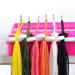 May include: A bright pink plastic loom tool featuring six vertical metal needles. Each needle holds a loop of embroidery thread in yellow, olive green, pink, orange, white, and black, with the threads hanging down in long, straight strands.