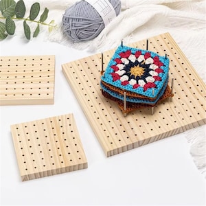 May include: Wooden crochet blocking boards with evenly spaced holes. A stack of colourful, square crochet pieces is pinned to one board. Additional smaller boards are also visible, along with a ball of grey yarn.