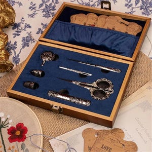 Puede incluir: Un costurero de madera con interior de terciopelo azul oscuro que contiene unas tijeras de metal ornamentadas, un dedal, un estuche para agujas, un pasacintas y un punzón. La tapa incluye un bolsillo con bobinas de hilo de cartón decorativas.