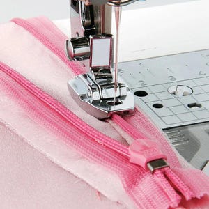 May include: A metal sewing machine presser foot is positioned over a pink zipper attached to pink fabric. The silver presser foot has a needle inserted through the center, and the sewing machine plate features a grid with visible measurement markings.