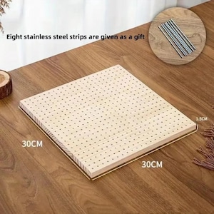 Può includere: Una tavola forata in legno chiaro, 30 cm x 30 cm e spessa 1,5 cm, con fori distanziati uniformemente. L'immagine mostra anche otto strisce di acciaio inossidabile. La tavola è su una superficie di legno.