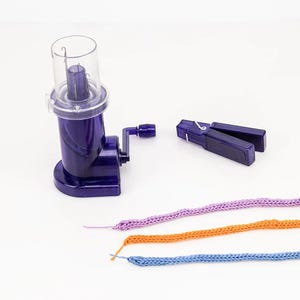 Può includere: Un avvolgitore di filato viola con una parte superiore trasparente, una manovella e un morsetto. Sono presenti anche tre cordoncini lavorati a maglia viola, arancioni e blu. Gli oggetti sono su una superficie bianca, suggerendo un tema artigianale.