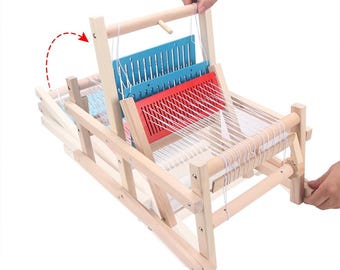 May include: A wooden weaving loom with red and blue plastic components. White thread is strung across the loom. The loom is being manipulated by a person's hands.