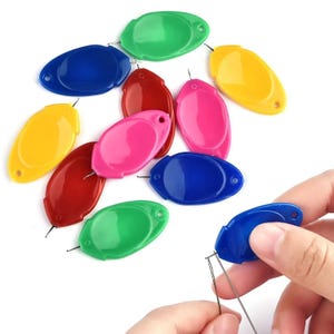 May include: A collection of oval-shaped plastic needle threaders in various colors including red, blue, green, yellow, and pink. Each threader features a small metal wire loop at one end and a hole at the opposite end for easy handling and storage.